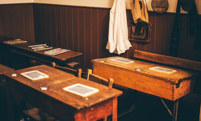 Victorian school desks with chalk writing boards on them. Victorian clothes hang on pegs in the background.